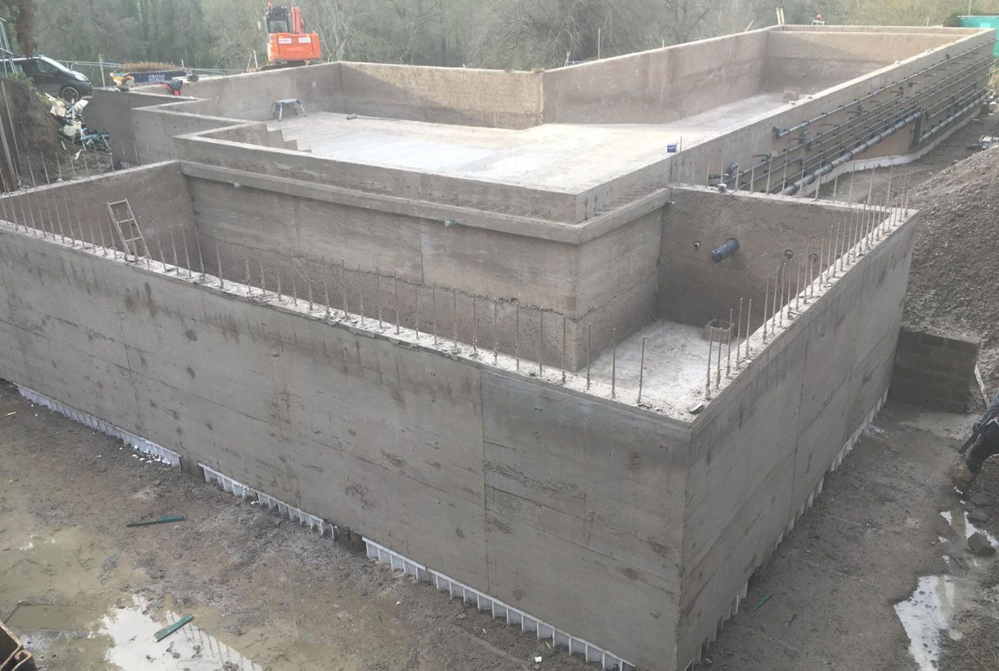 Concrete foundation of an infinity pool shell under construction on a muddy site, with a worker visible in the background.