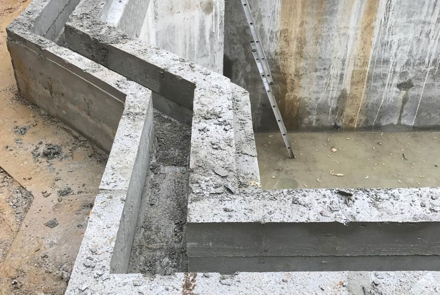 Detail of a concrete swimming pool shell with overflow channel with fresh cement and a muddy construction site.