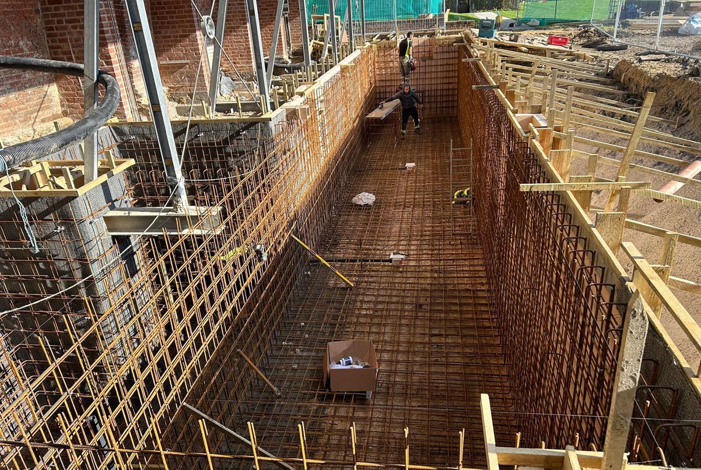 Rectangular swimming pool under construction with steel rebar walls, wooden formwork and workers at the far end.