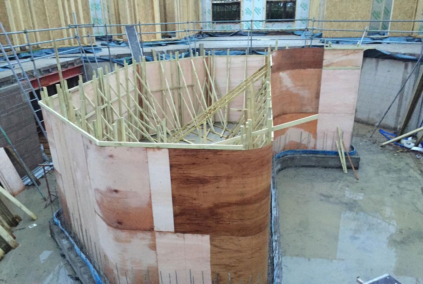 Curved wooden formwork for a swimming pool with vertical supports at an indoor construction site, surrounded by scaffolding.