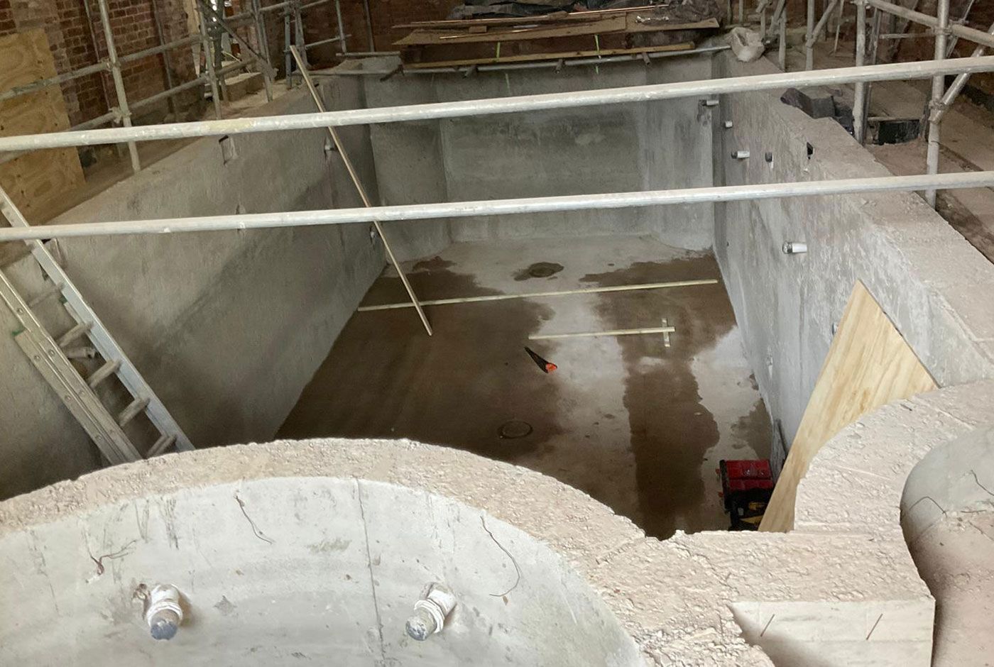 Indoor swimming pool concrete shell under construction with a ladder, scaffolding and base drying off.
