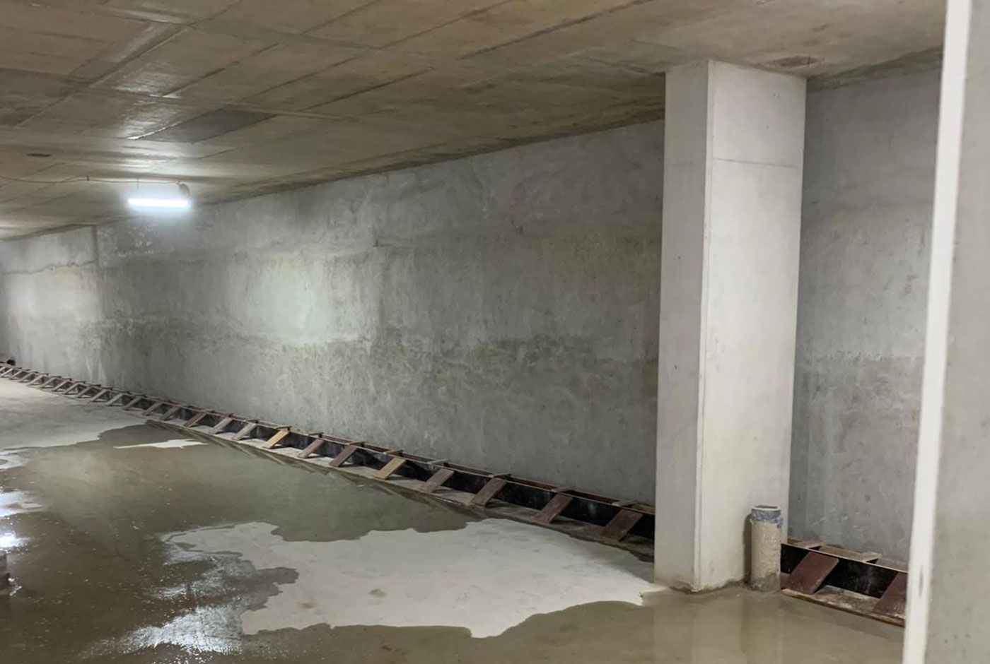 Large underground space with concrete walls, floor and ceiling, a bright light shining on the wet floor.