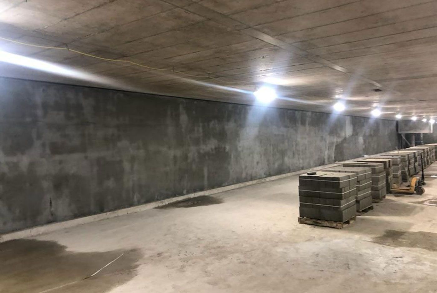 Industrial space with concrete walls and a row of ceiling lights, stacks of materials lined along the right side.