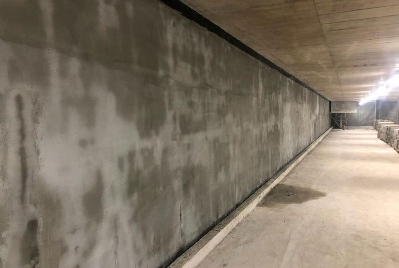 Interior space with a long grey concrete wall lit by a string of ceiling lights.