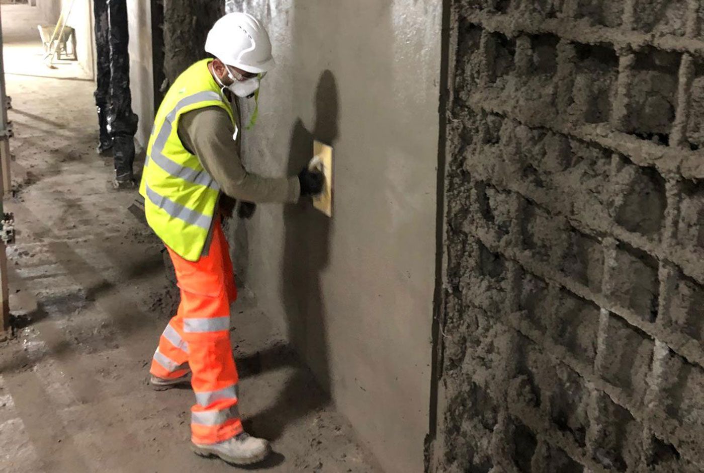 Construction worker in hi-viz kit, hard hat and mask smoothing freshly applied concrete on a section of wall.