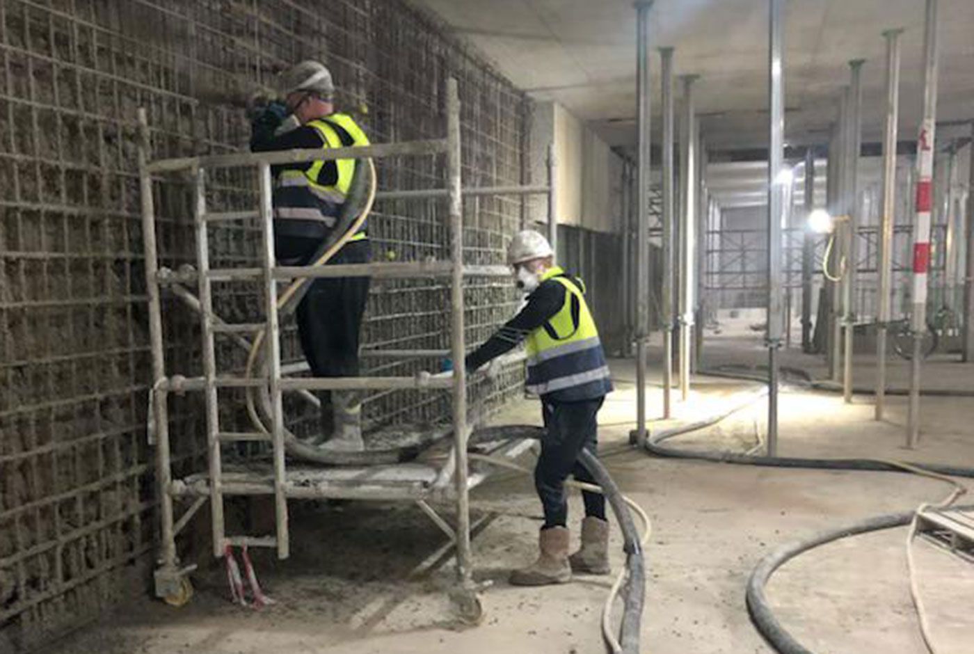 Workers in hi-viz and hard hats on a low scaffolding platform, one spraying concrete onto a wall covered with a metal grid.