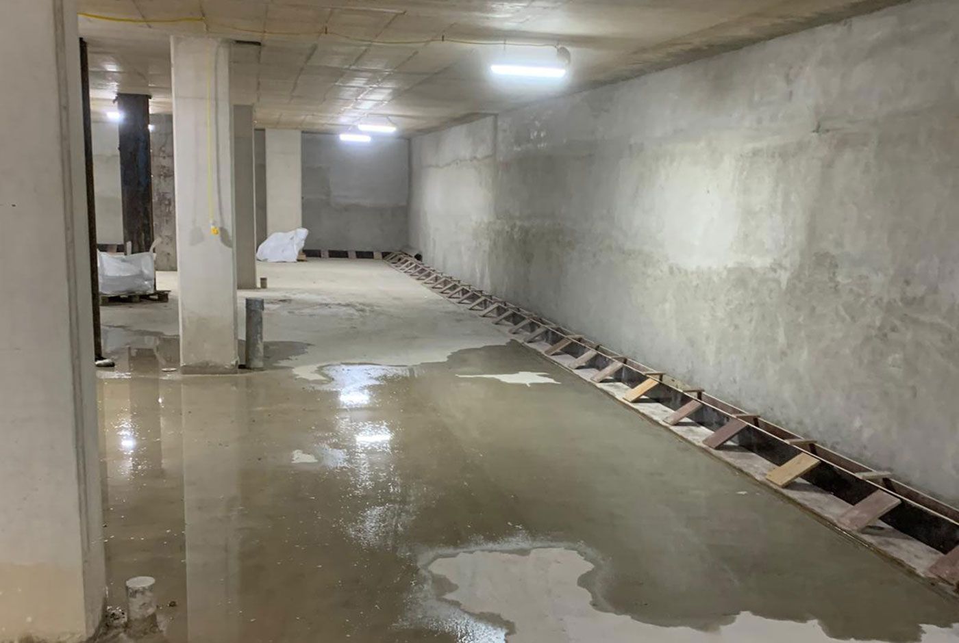 A concrete basement or car park with standing water on the floor lit by ceiling lights.