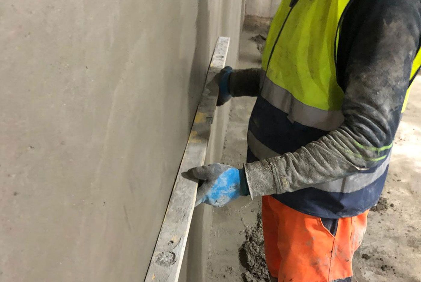 Construction worker in a hi-viz vest and blue gloves levelling sprayed concrete on a wall.