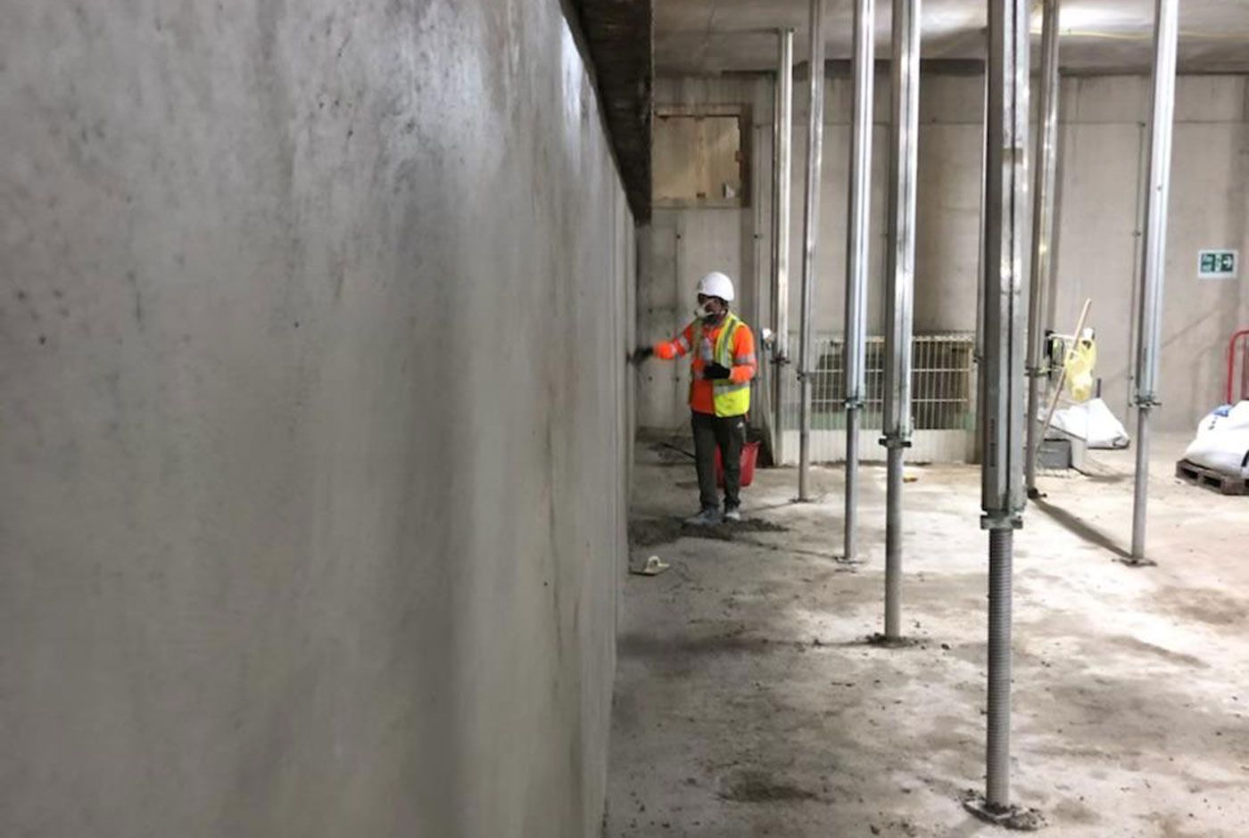 Construction worker in hi-viz and hard hat smoothing freshly applied concrete on a wall, with metal ceiling supports.