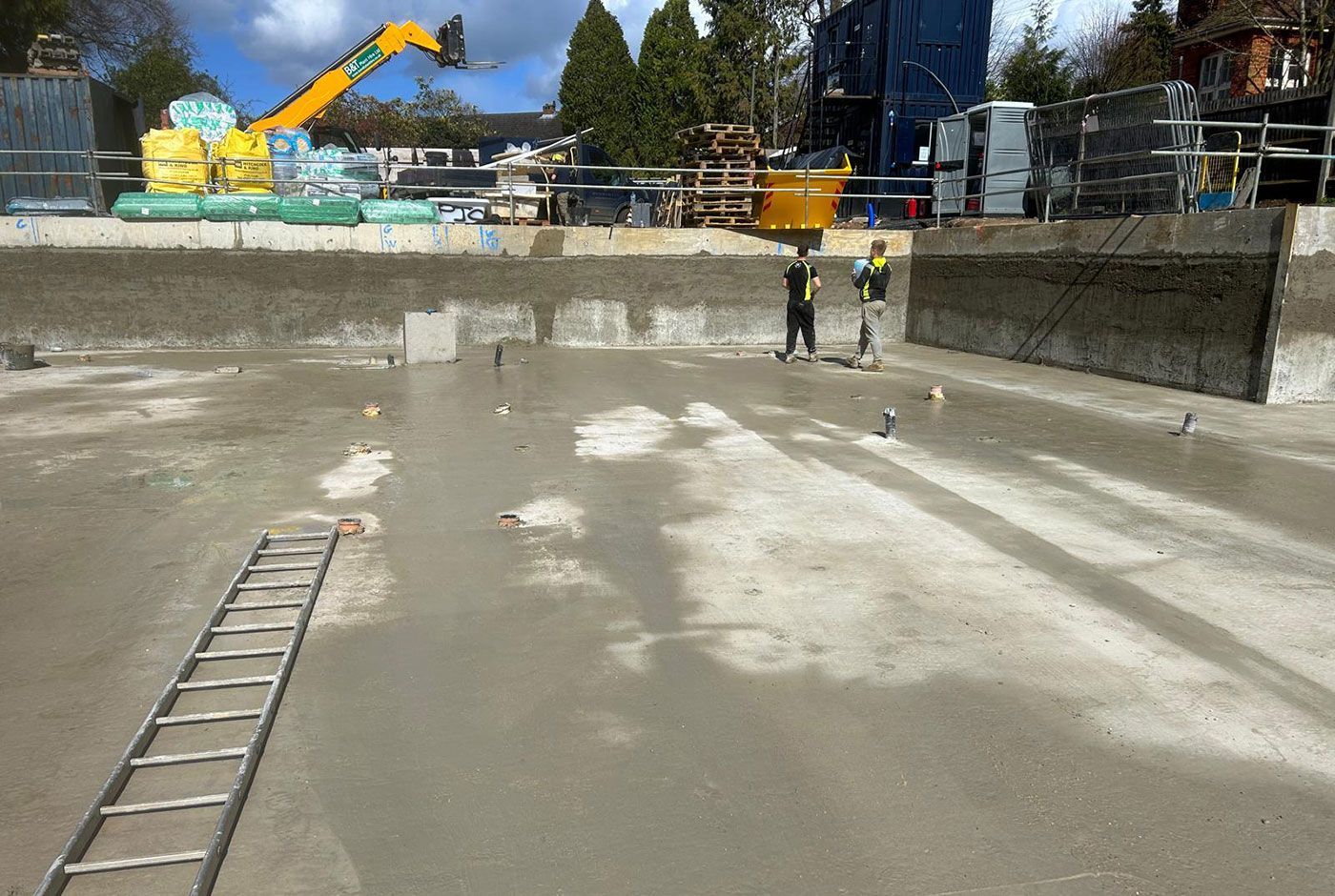 Construction site with concrete basin, ladder in foreground, and excavator in the background.