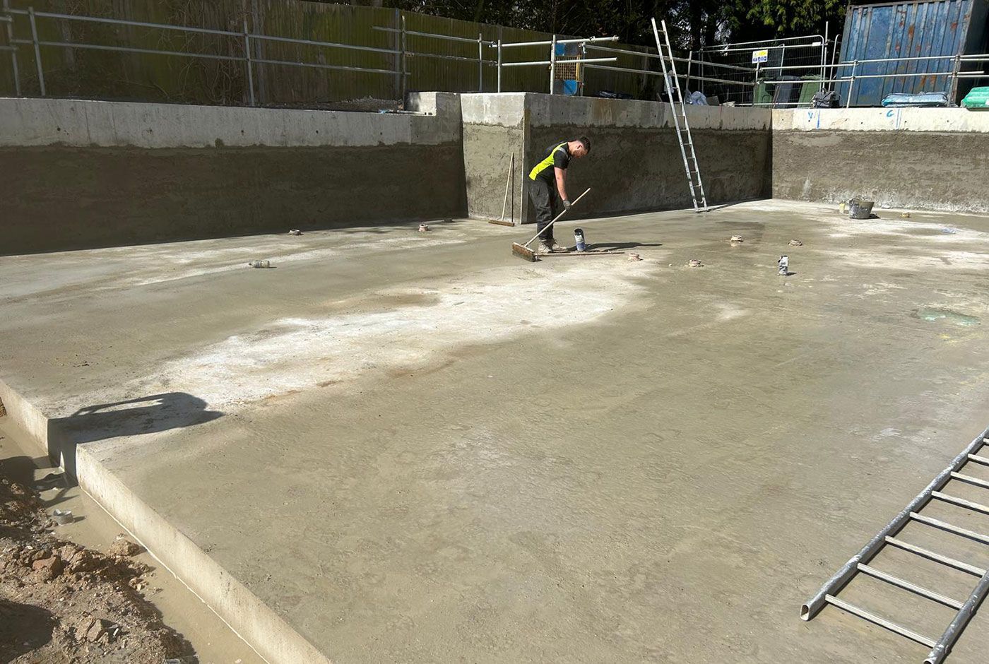 Construction worker smoothing a wet concrete base with shallow walls and several ladders visible.