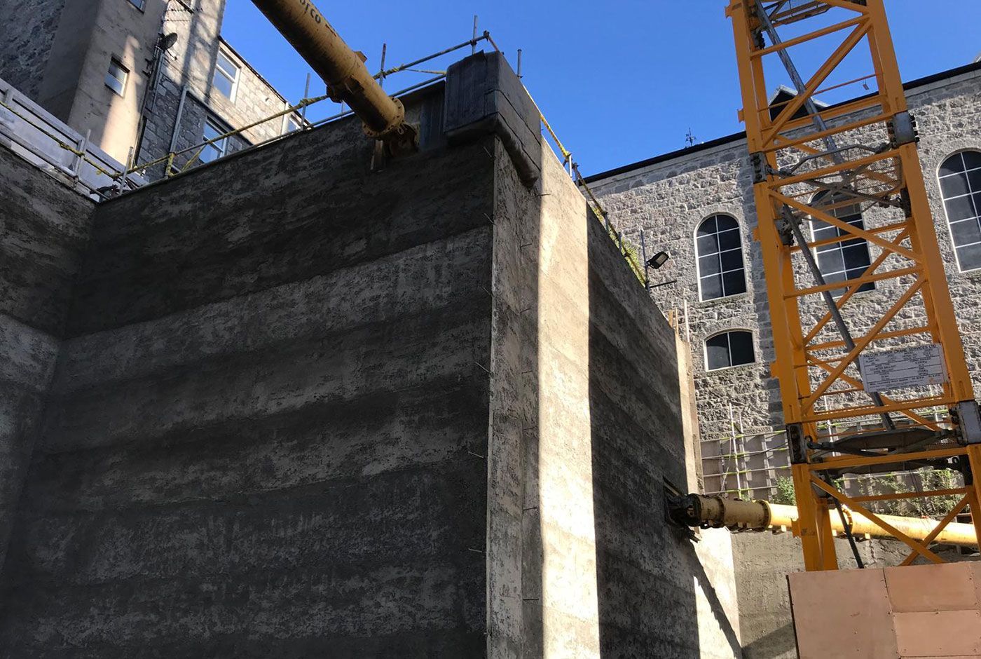 Construction site with concrete walls and yellow crane against a bright blue sky, old building in the background.