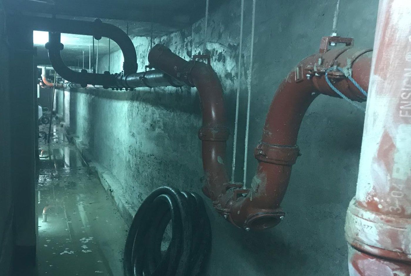 Industrial pipes and valves in a dimly lit, wet basement or utility room.