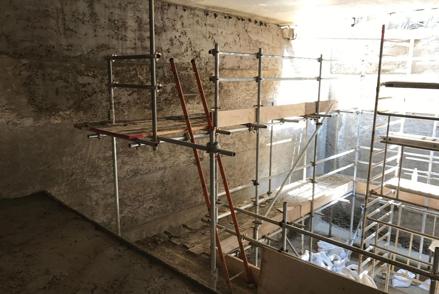 Interior construction site with rough concrete walls, scaffolding, work platforms and ladder, bright sunlight from above.