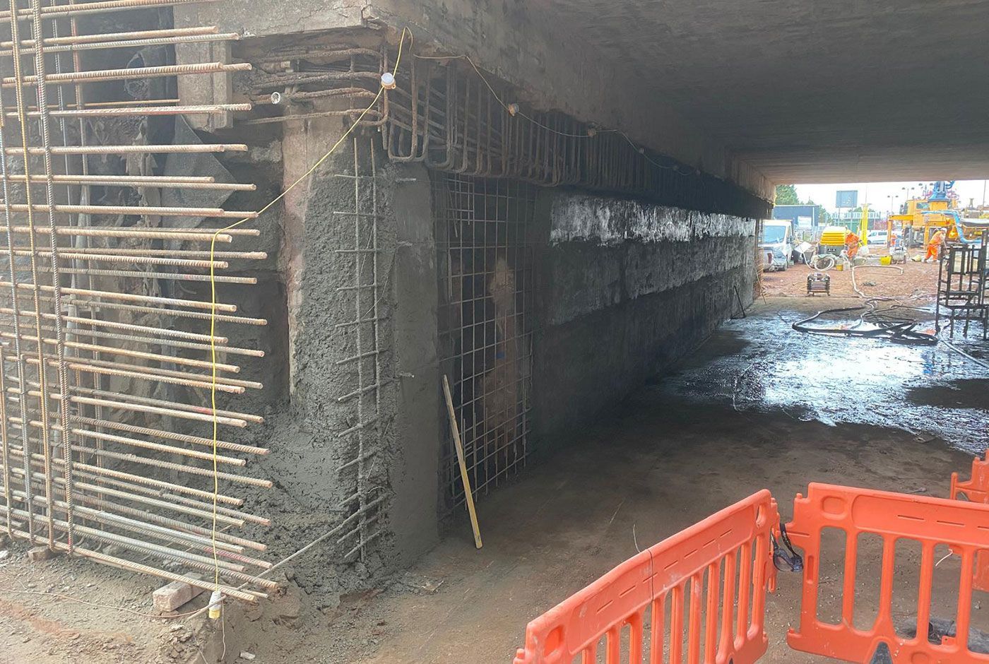 Pile facing being carried out under a bridge with steel rebar forms, safety barriers, construction work in the background.