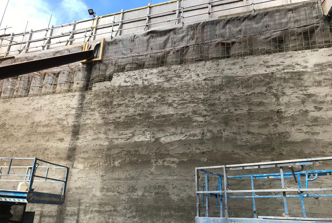 Pile facing a retaining wall under construction with two scissor lifts against a blue sky.