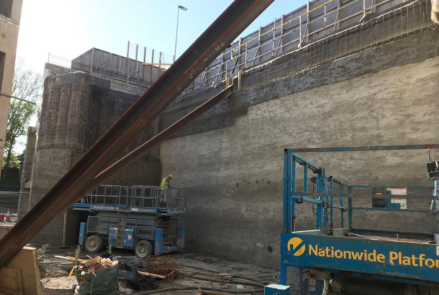 Pile facing a concrete wall on a construction site, with workers and two blue scissor lifts.