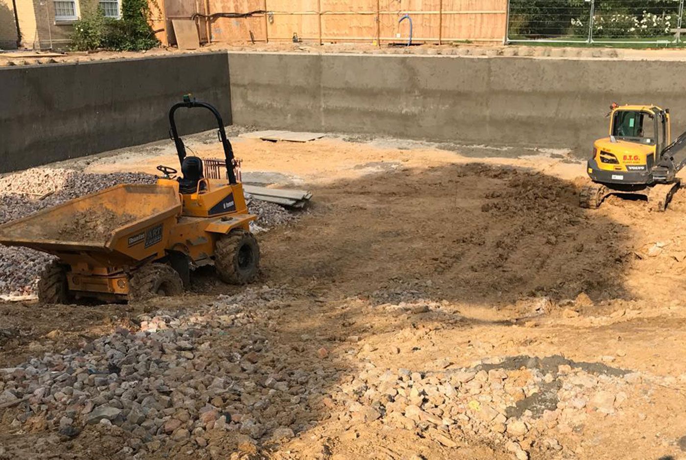Two yellow construction vehicles in a foundation pit with sand and rock, concrete walls in the background.