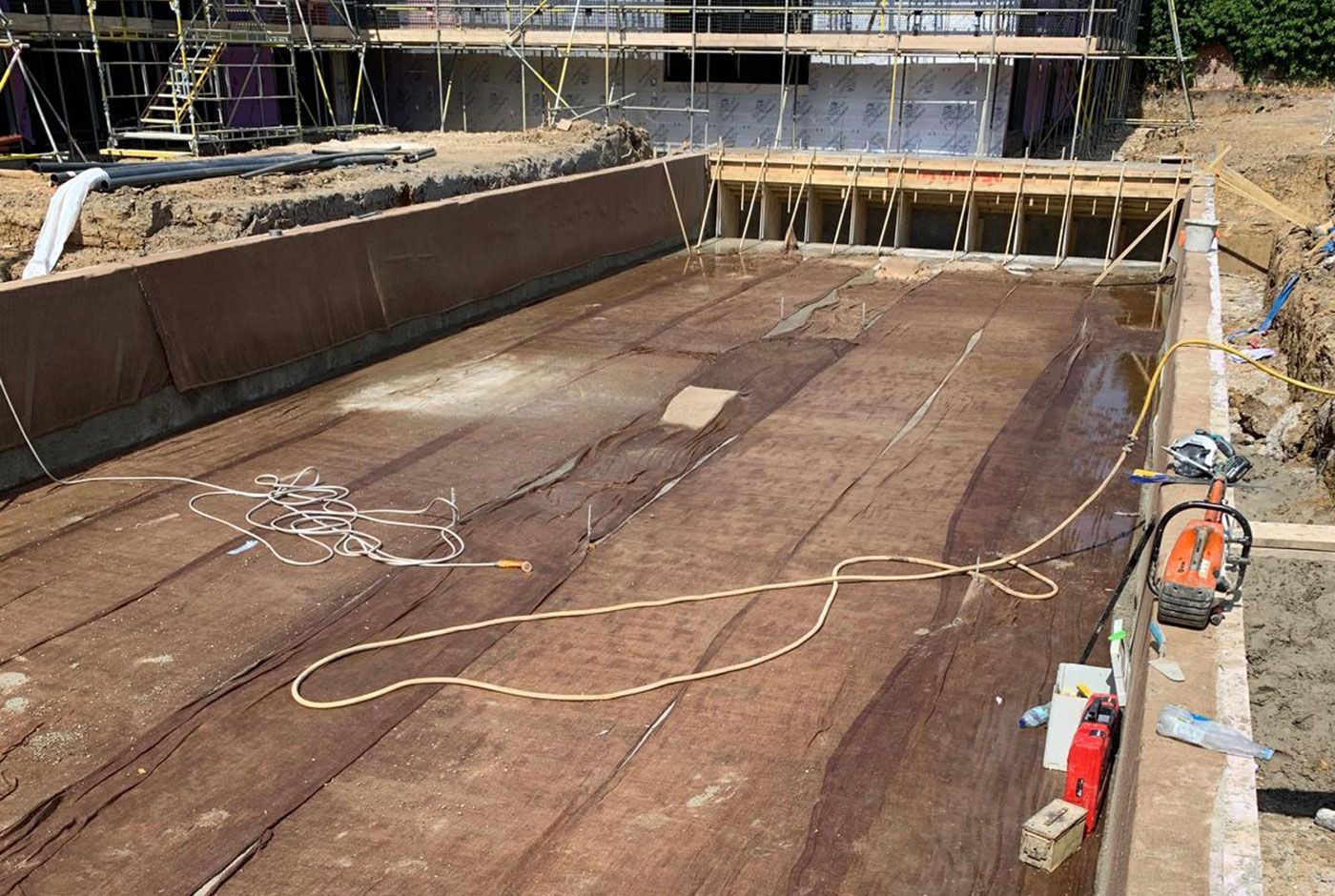 Large exterior skimmer pool under construction, with boarded framework, the wider construction site surrounding.