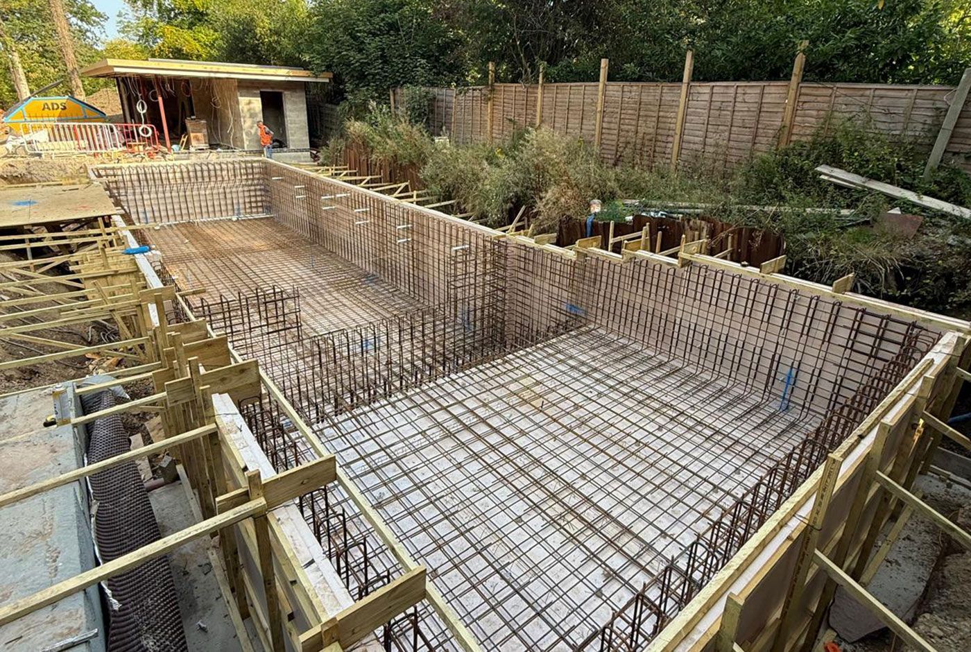 Rebar and wooden formwork for the concrete shell of a skimmer pool, with shrubbery and fencing in the background.