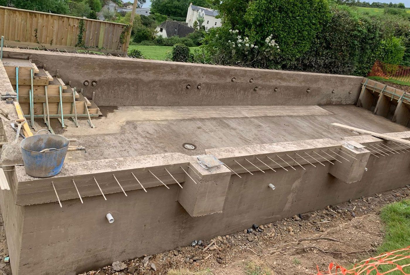 The shell of a skimmer pool under construction in a country garden, with exposed rebar and unfinished walls and steps.