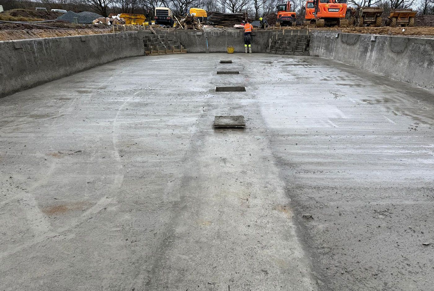 Low level view of a skimmer pool shell, with rebar supports, a construction worker and several excavators in the background.