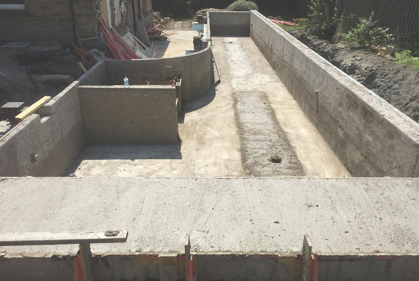 An outdoor skimmer pool under construction in a residential garden, with concrete walls and part of the base is still drying.