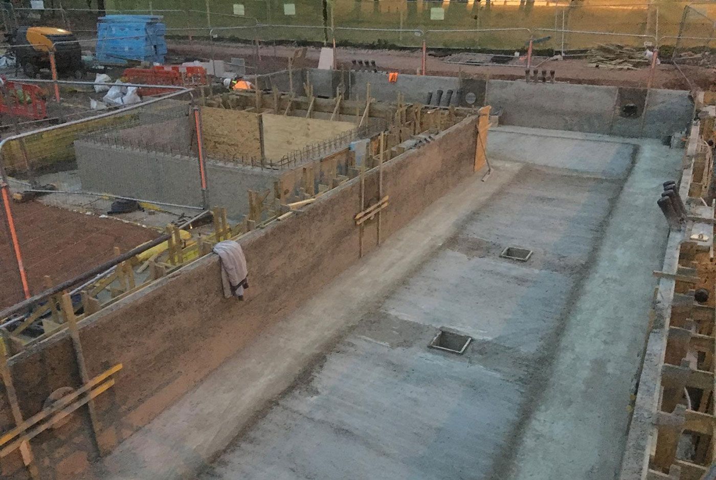 Skimmer pool construction site with concrete base, wooden forms, surrounded by safety fencing and construction materials.