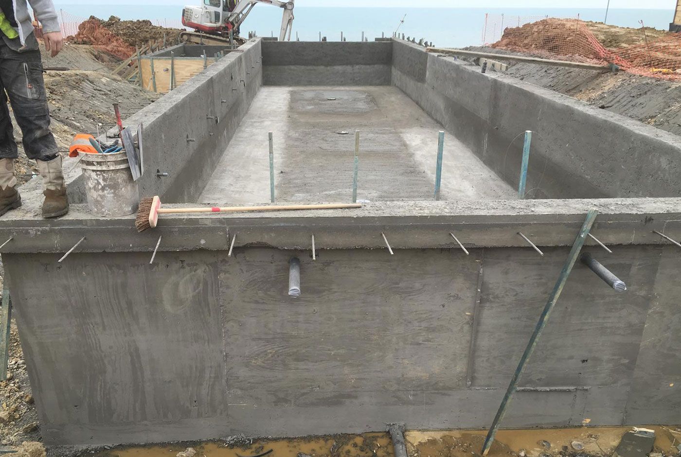 Concrete skimmer pool shell under construction at a coastal location, with wooden forms and mini excavator in the background.