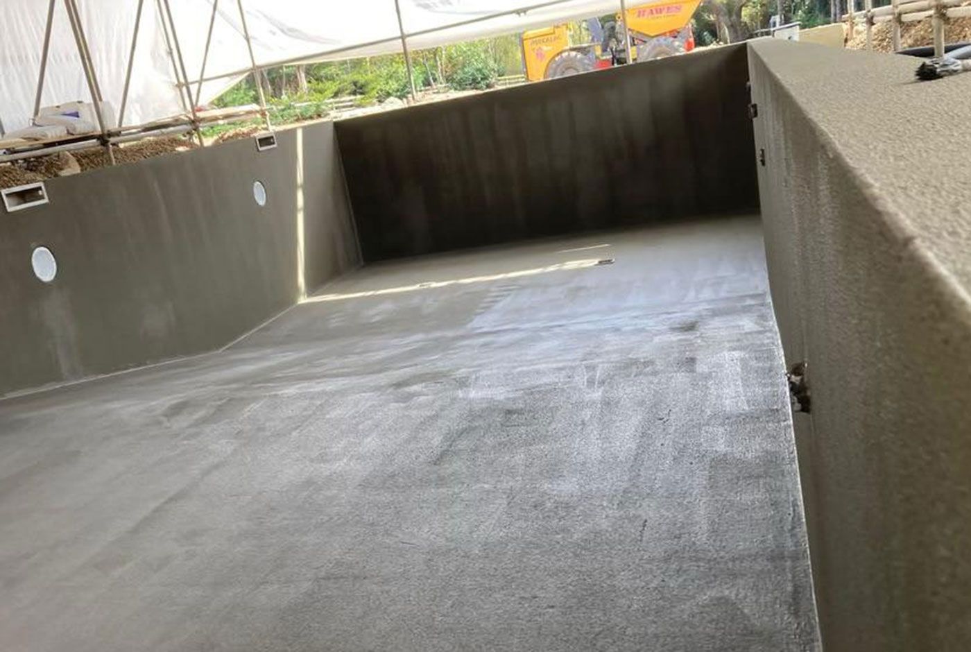 Concrete shell of an outdoor skimmer pool basin drying under a canopy, viewed from one end.