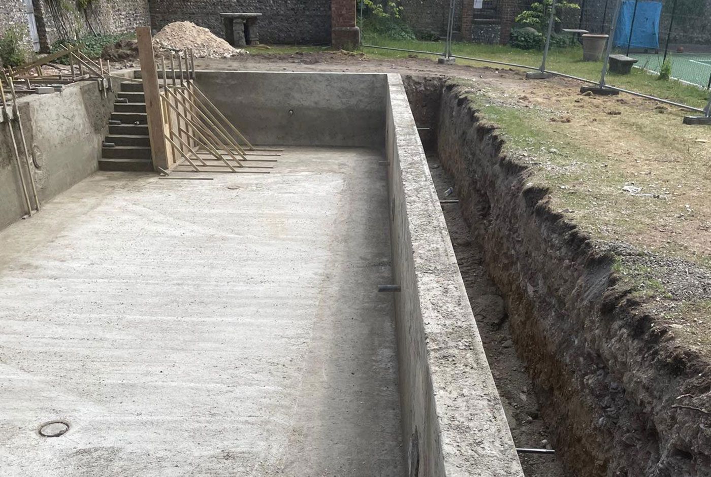 A skimmer pool shell constructed adjacent to a property, with steps just formed from concrete and the area fenced off.