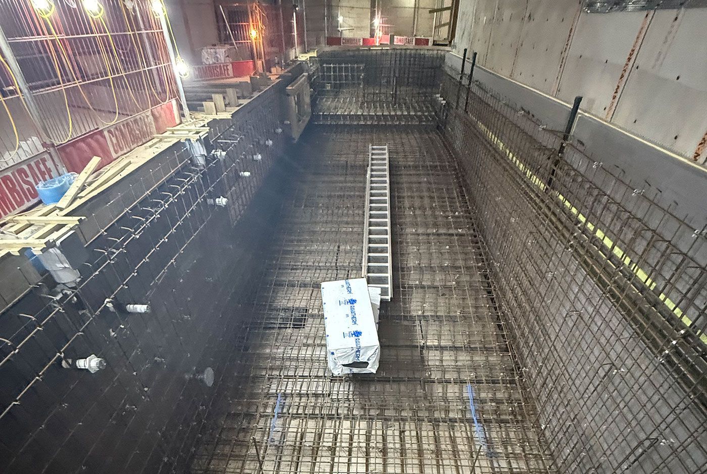 A skimmer pool under construction, with steel rebar walls and mesh framework, a ladder on the base, under lights.