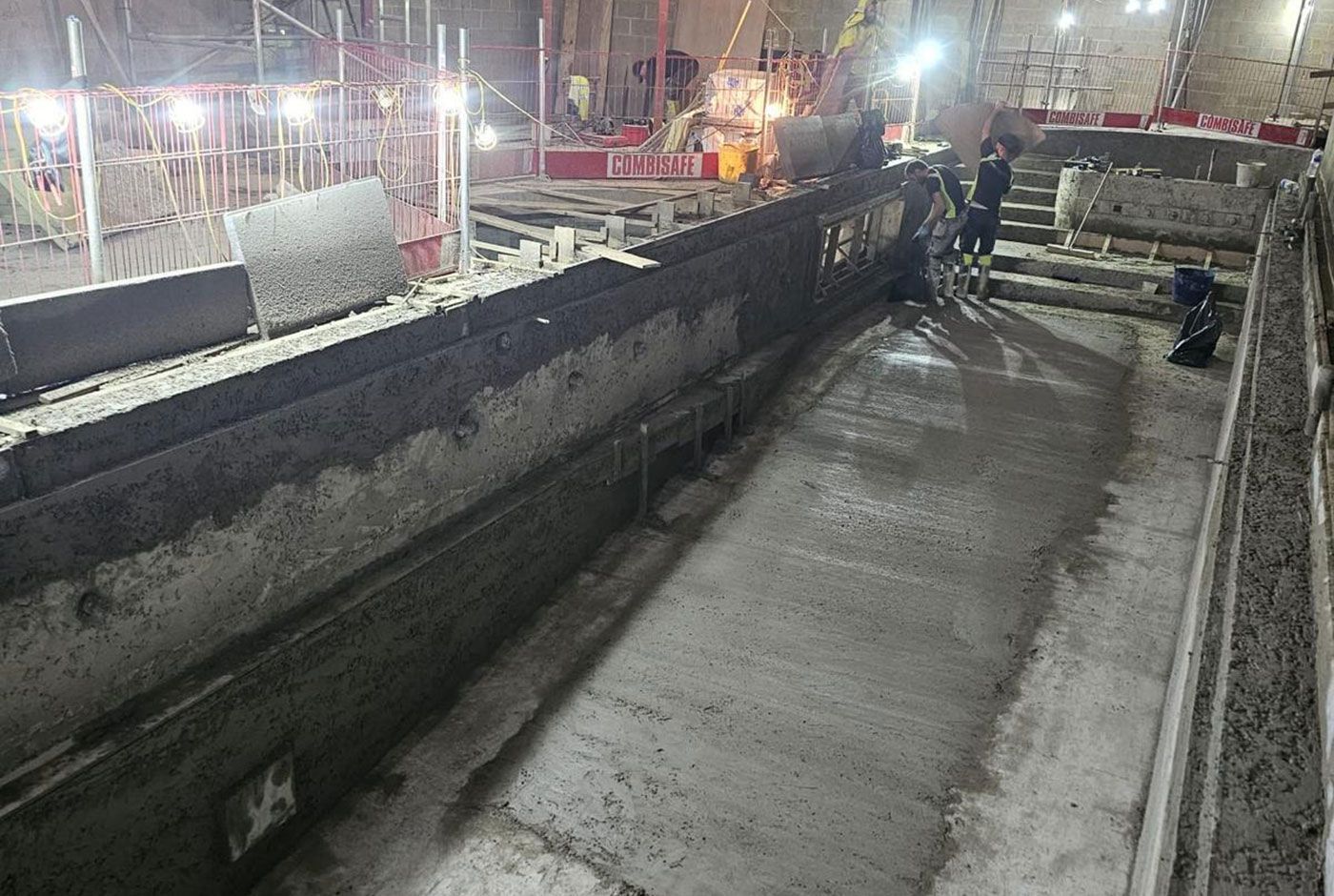 Indoors construction site with work on a skimmer pool shell taking place under floodlights. 