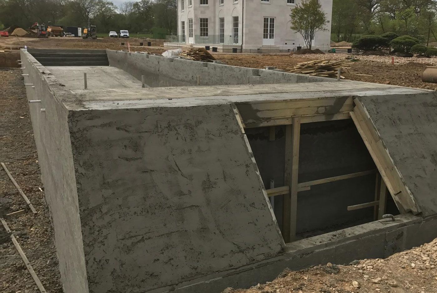 Concrete skimmer pool shell under construction in the grounds of a large house,  the surrounding lawns have been removed.