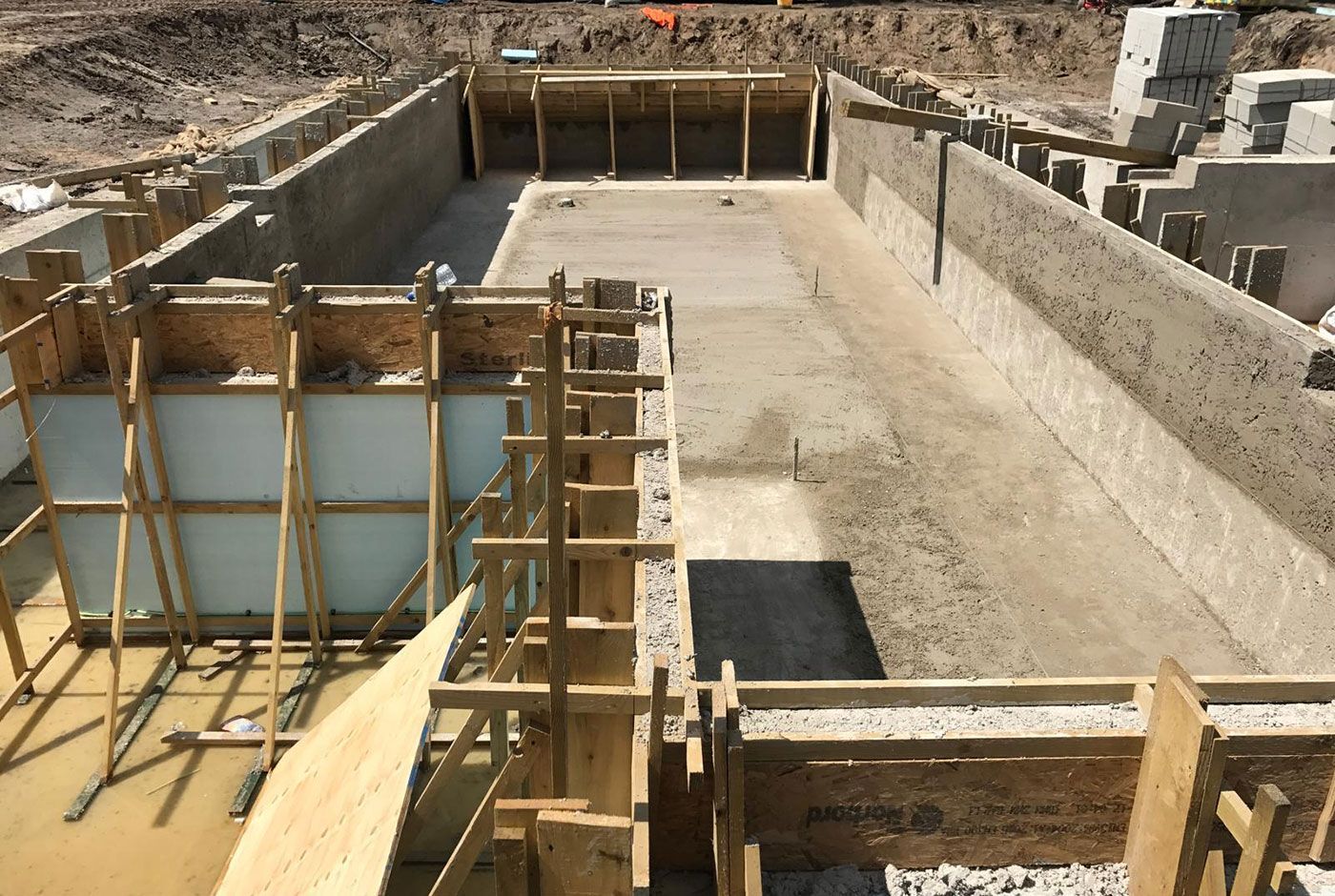 Construction site with concrete walls and base of a skimmer pool, with wooden supports, surrounded by building materials.
