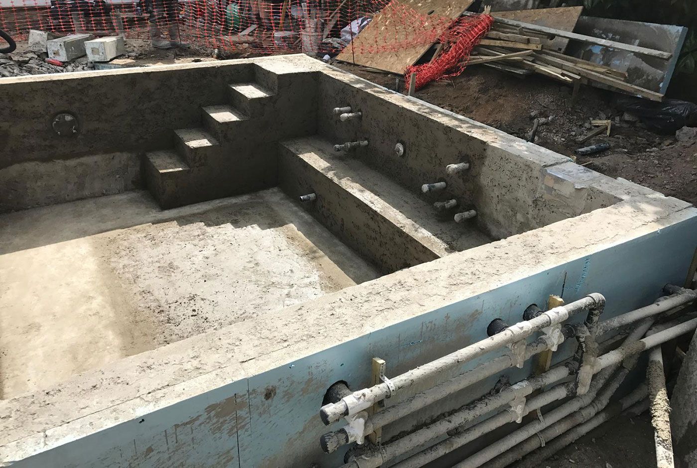 Concrete rectangular pool shell under construction with steps and exposed pipes, orange fencing and materials in background.