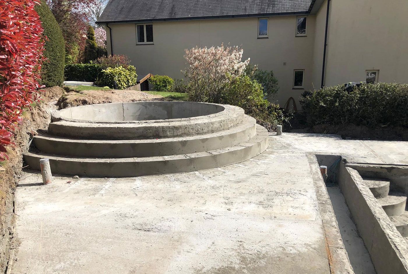 Stepped circular concrete shell of a spa pool in a sunlit courtyard with shrubs and building in the background.