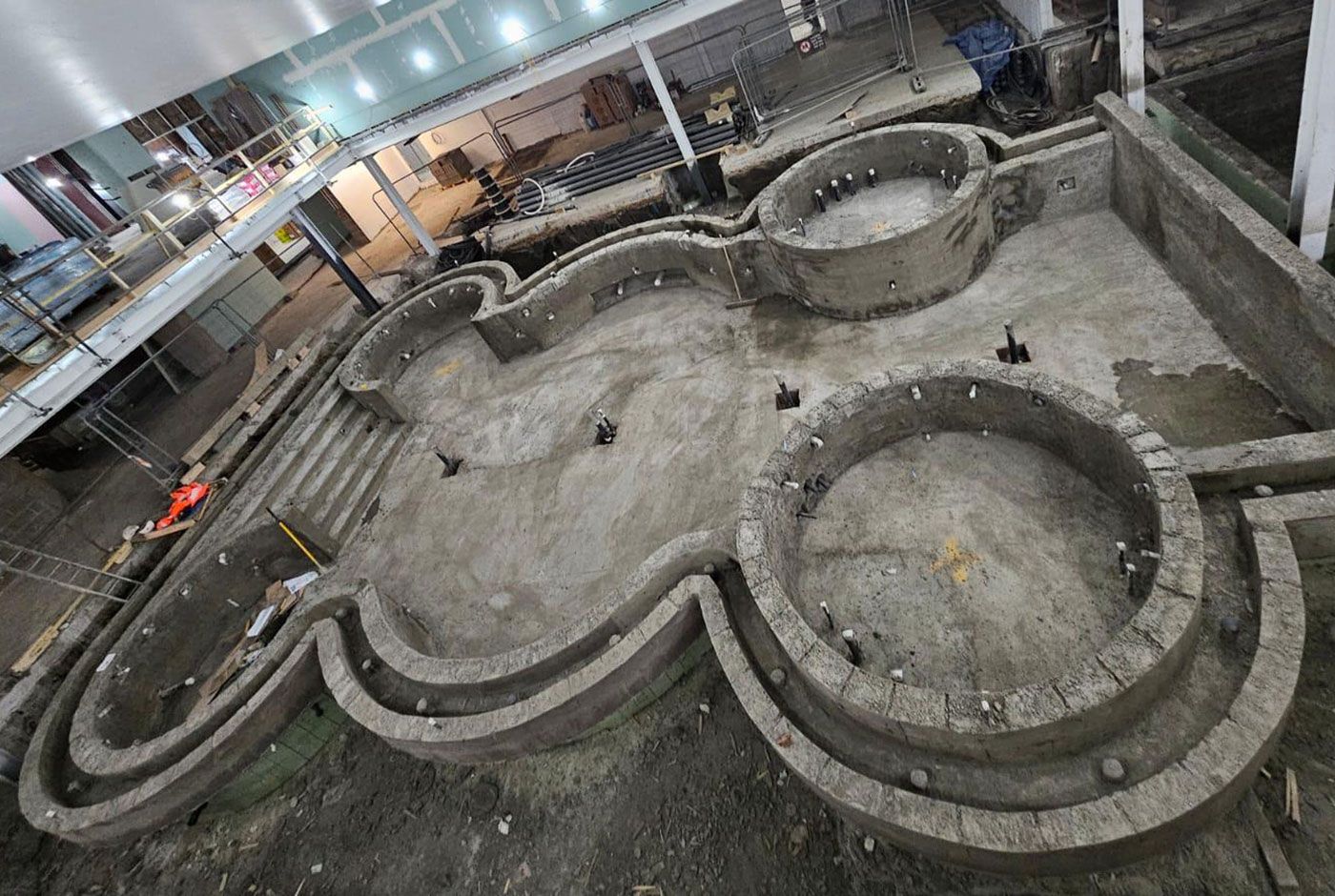 Overhead view of the concrete shell of a large indoor pool with two circular spa areas.