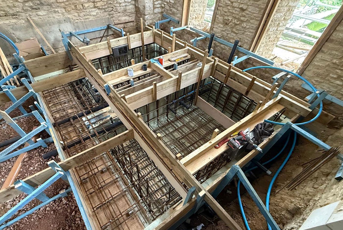 Construction site for a plunge pool with  rebar grid, wooden formwork, and blue support struts inside a bare brick building.