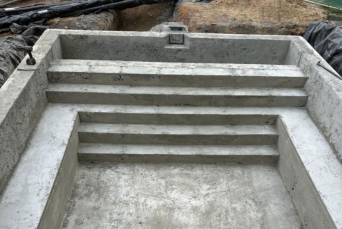 Concrete shell of an outdoor rectangular pool with steps and seating, surrounded by excavated ground and black plastic.