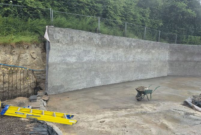 Concrete retaining wall under construction with a wheelbarrow and collapsed step ladders, wooded hillside in the background.