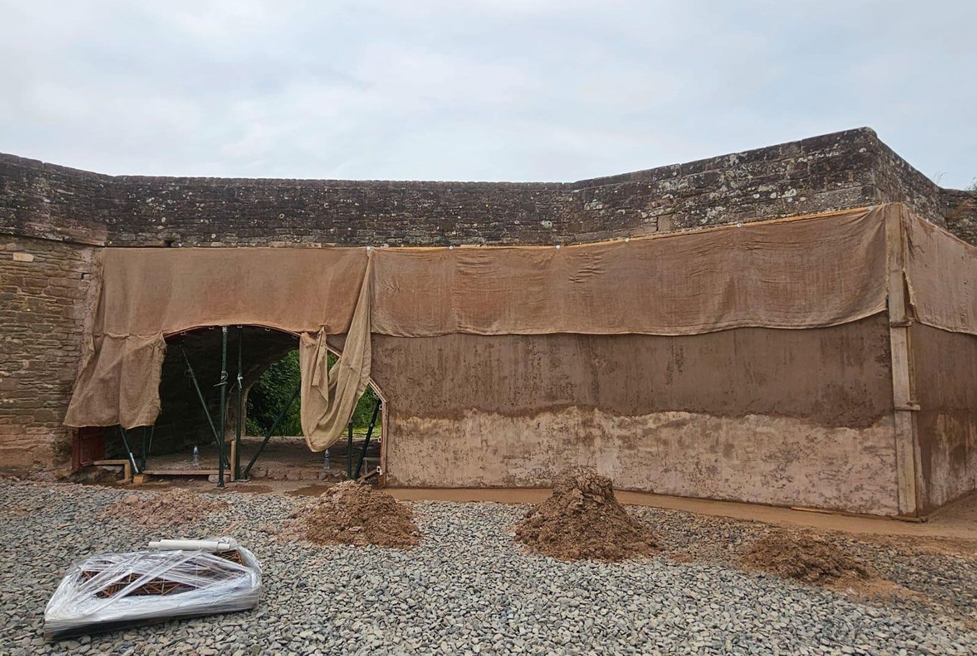 An old stone bridge with sprayed concrete applied to the structure, material draped over the top, gravel in the foreground.