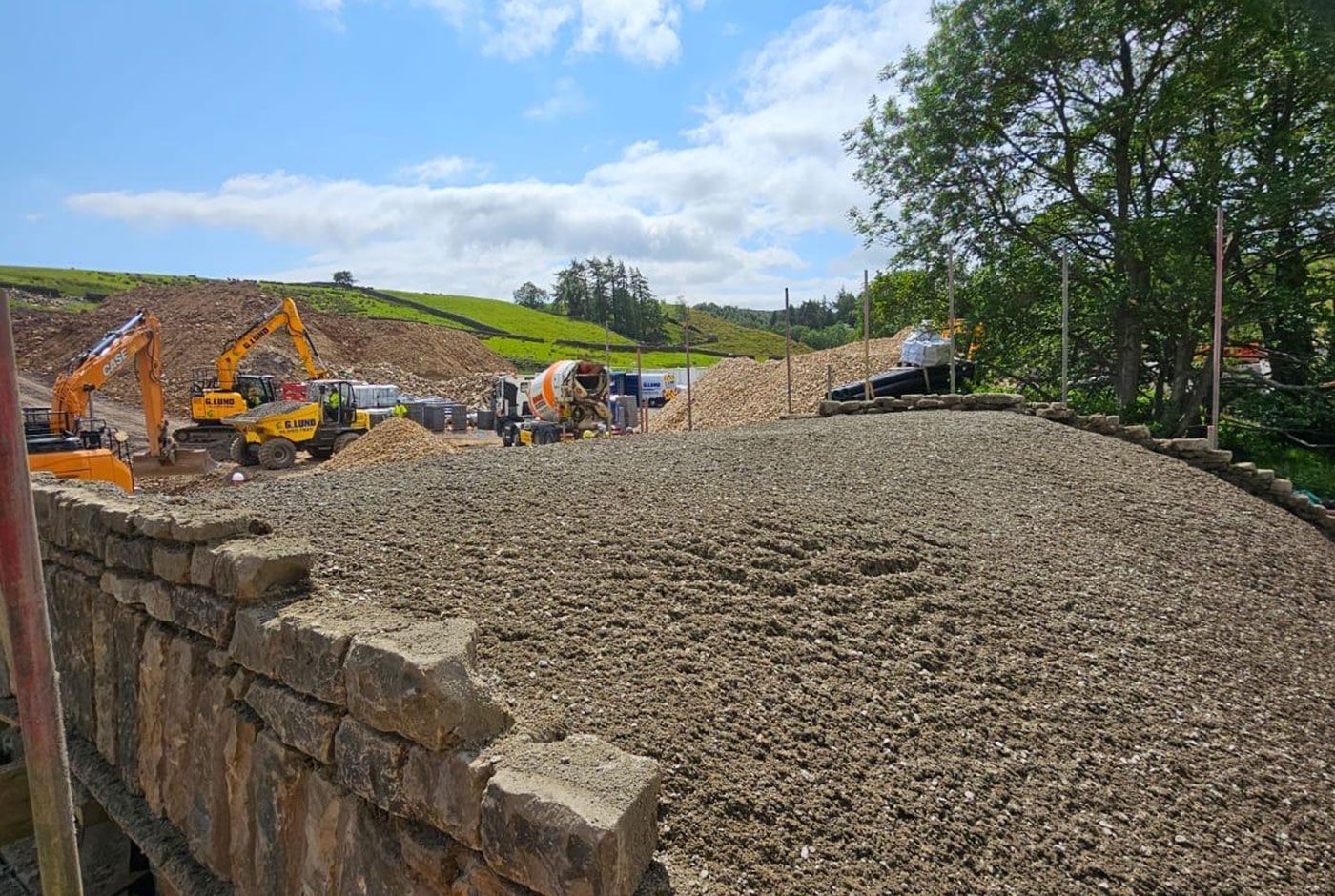 Construction site in a rural location with excavators and piles of gravel beside a stone wall under a blue sky.