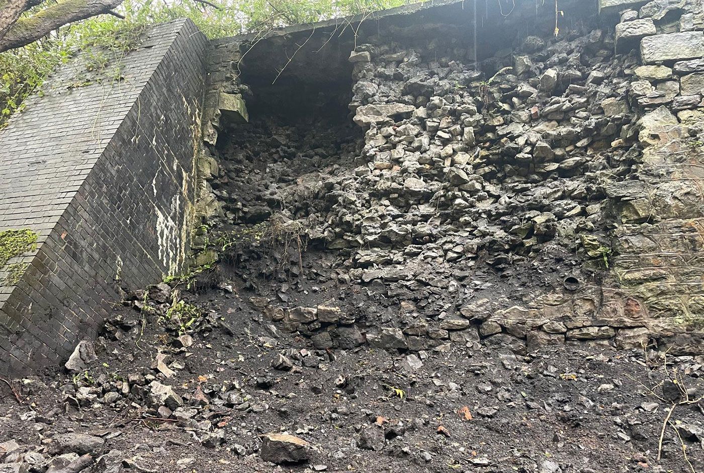 A old stone retaining wall that has collapsed with loose materials next to a buttress.