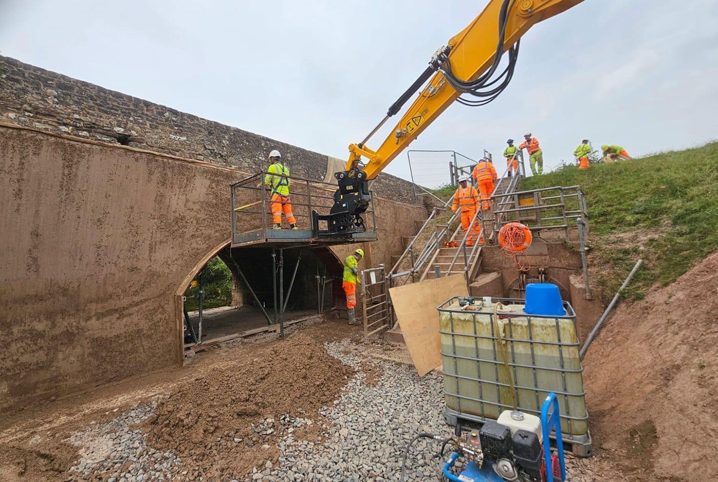 Construction workers using an excavator near a tunnel entrance on a muddy, sloped work site