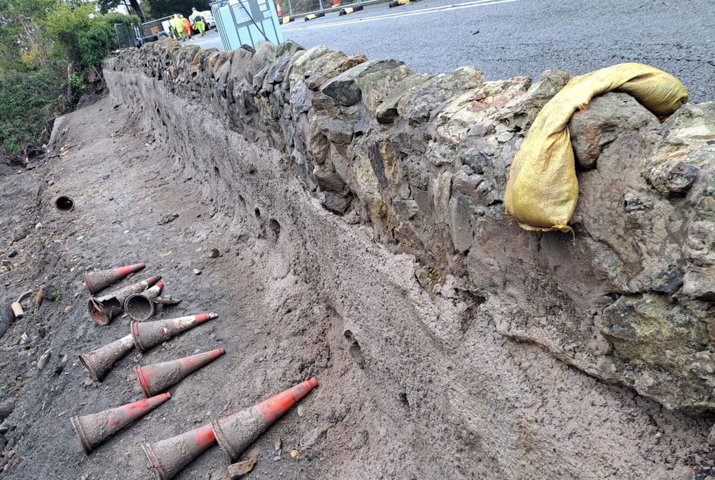Stone wall repair with tools and sandbag, muddy red cones laid on a dirt slope beside a road.