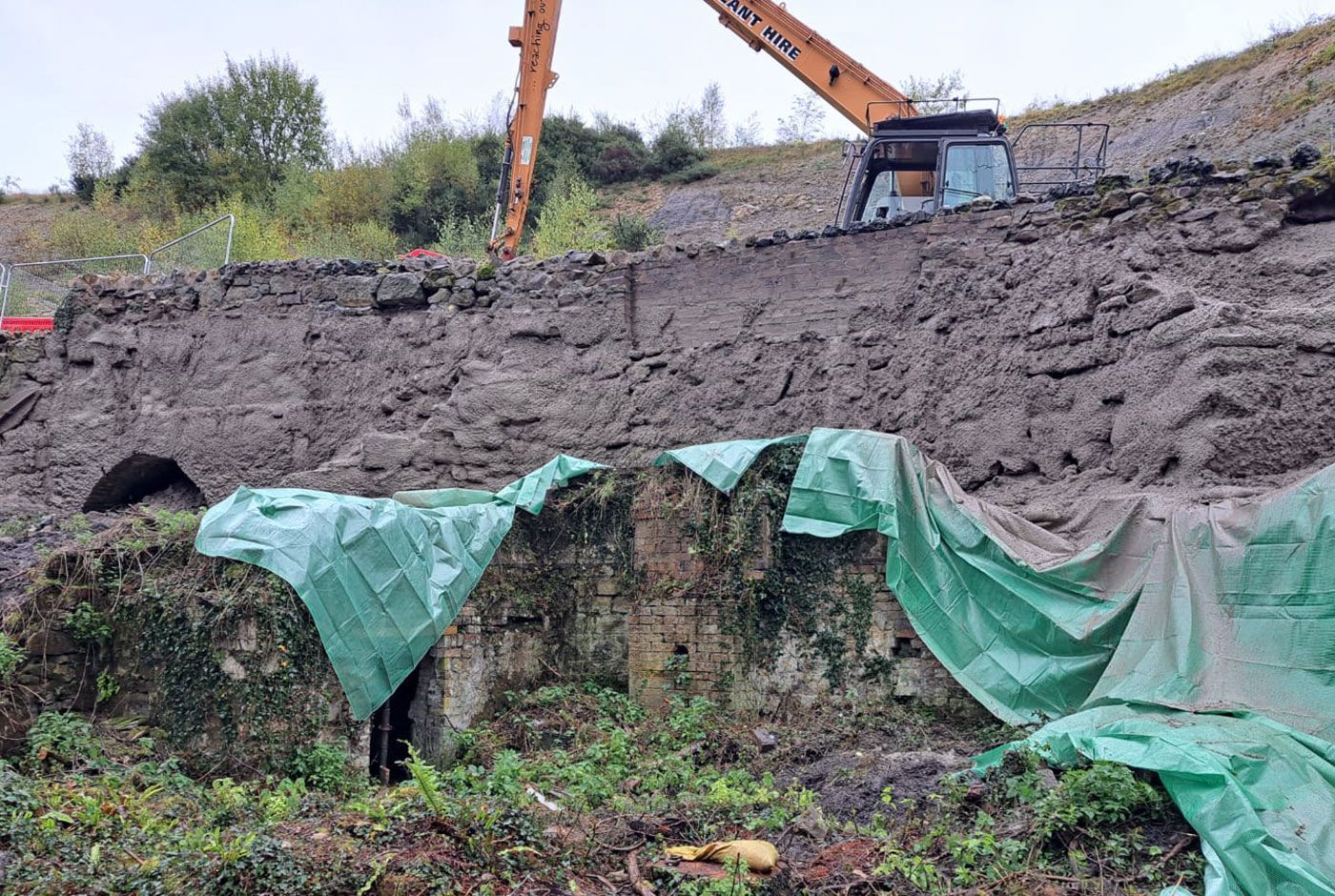 Excavator on a road bridge under repair, its retaining wall roughly covered with concrete, green tarpaulins draped..