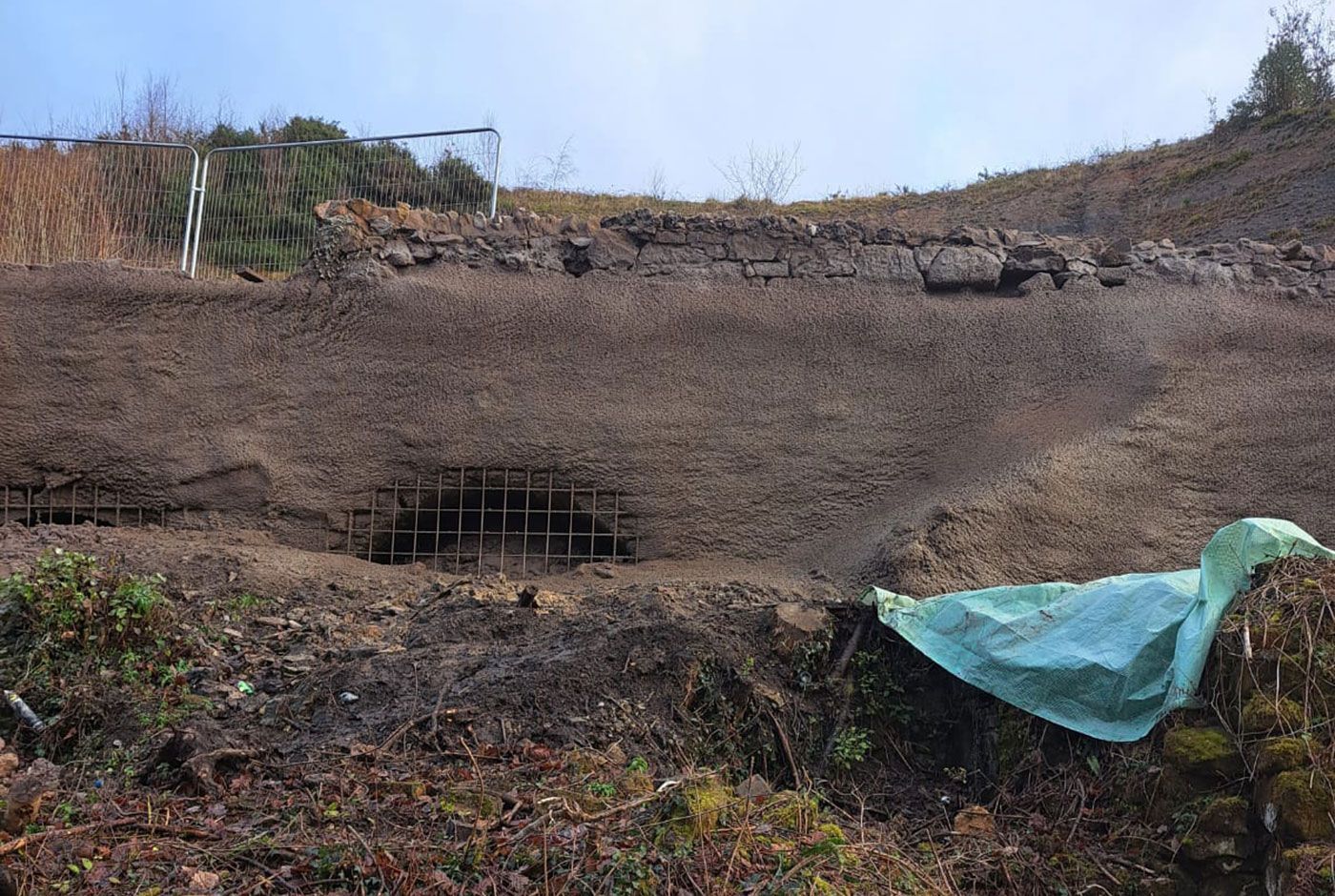 A retaining wall under repair with concrete applied, rocky ground, and a turquoise tarpaulin on the right