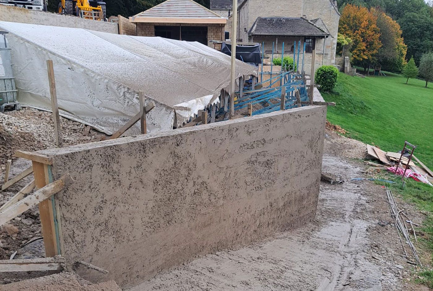 Large unfinished concrete retaining wall beside a house, with building materials on a muddy site, lawn beyond.