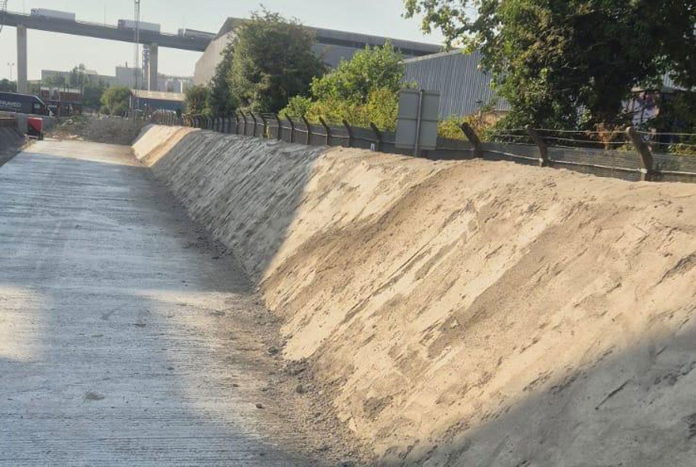 Sloping concrete retaining wall, with trees, fencing, and a bridge in the background.